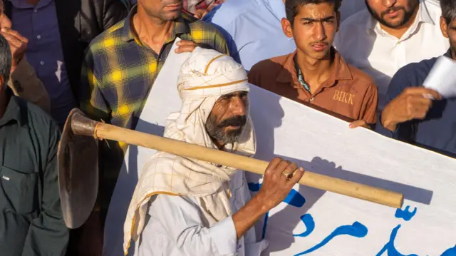 Beyaz başlık ve beyaz kıyafetler giymiş bir adam, omzunda bir kazma taşırken arkasında diğer protestocular görülüyor.