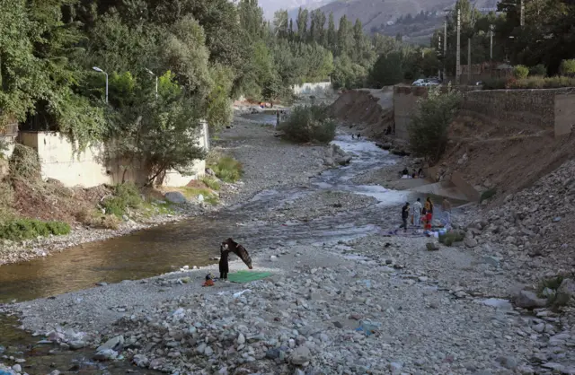 İnsanlar quru, daşlı çay yatağında xalçanın üzərində oturublar. Çayın ortasından dar bir kanal keçir və oradan çox az su axır.