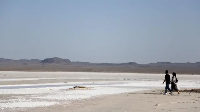 Bir erkek ve bir kadın, yüzeyinde tuz birikintileri bulunan, dağların uzakta göründüğü kurumuş Hoz-e Soltan Gölü yatağında yürüyor. Qum şəhərindəki Hoz-e Soltan duz gölü demək olar ki, tamamilə quruyub.